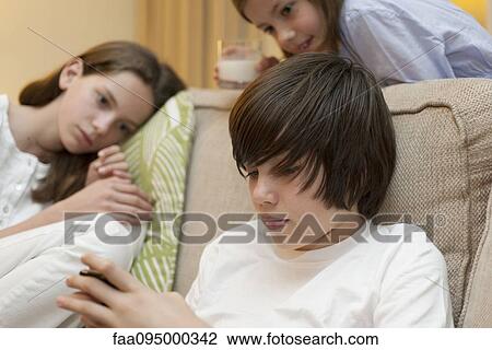 Stock Image - Teenage boy using smartphone while sisters watch in background. Fotosearch