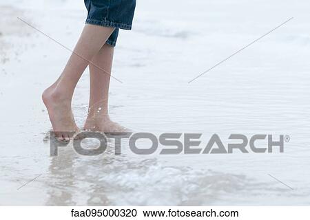 Stock Image - Teenage boy wading in surf at the beach, low section. Fotosearch