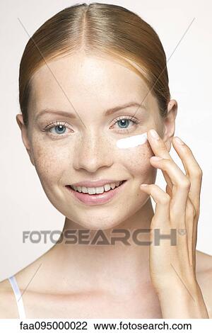 Stock Image - Young woman applying moisturizer under eye. Fotosearch