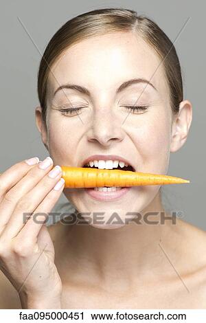 Stock Image - Young woman biting into carrot, eyes closed. Fotosearch