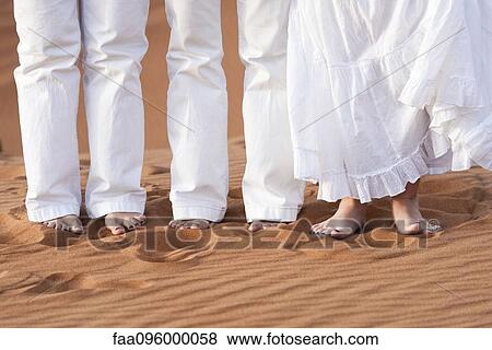 Stock Photo - Family wearing white clothing, standing barefoot in sand, low section. Fotosearch