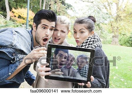 Stock Image - Friends photographing themselves making funny faces with digital tablet outdoors. Fotosearch