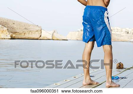 Stock Image - Teenage boy in swimming trunks standing on dock. Fotosearch