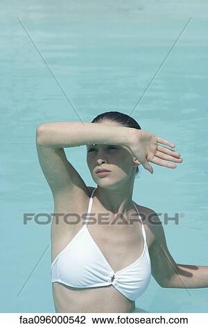 Stock Image - Young woman in swimming pool shielding eyes. Fotosearch