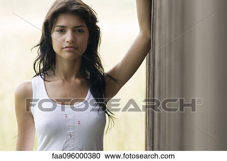 Stock Image - Young woman leaning against wall, looking at camera, portrait. Fotosearch