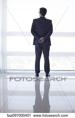 Stock Image - Businessman looking out high rise window at view of city below, rear view. Fotosearch