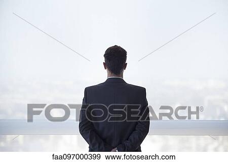 Stock Photo - Businessman looking out high rise window at view of city below, rear view. Fotosearch