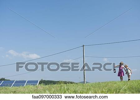 Stock Image - Children walking in field near solar panels and power lines, carrying old-fashioned lantern. Fotosearch