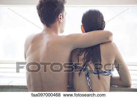 Stock Photo - Couple at edge of indoor pool looking out window at view, rear view. Fotosearch