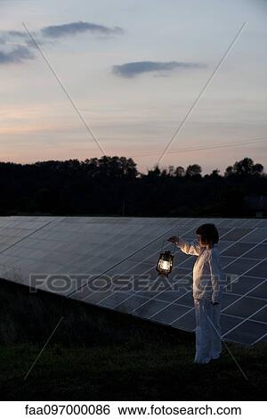 Stock Photograph - Girl standing in front of solar panels at twilight with old-fashioned lantern in hand. Fotosearch