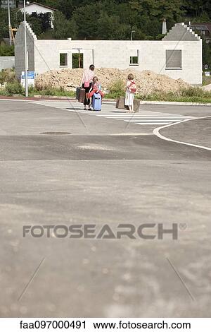 Stock Image - Mother and children walking in street toward unfinished house, dragging suitcases behind them. Fotosearch