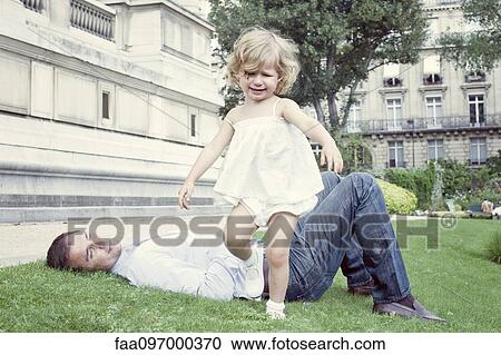 Stock Image - Toddler girl walking outdoors, father lying on grass in background. Fotosearch