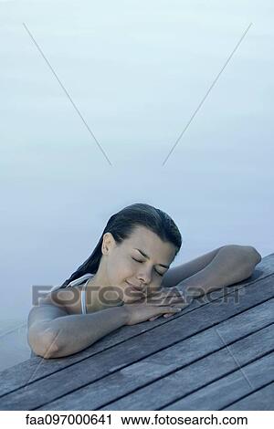 Stock Image - Woman in pool leaning against deck, resting head on arms, eyes closed. Fotosearch
