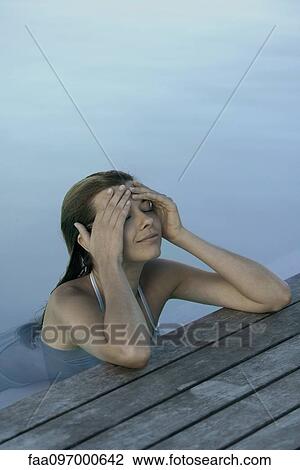 Stock Image - Woman in pool leaning against deck, holding head, smiling. Fotosearch