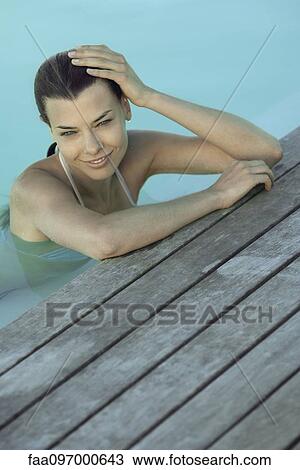Stock Image - Woman in pool leaning against deck, holding head, smiling. Fotosearch
