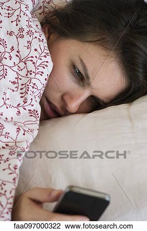 Stock Image - Woman lying bed looking at smartphone with unhappy expression on face. Fotosearch