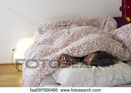 Stock Photography - Woman lying in bed under covers, looking at smartphone. Fotosearch