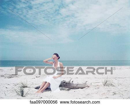 Stock Photograph - Woman sitting on driftwood at the beach, hand covering eyes. Fotosearch