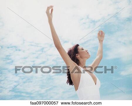 Stock Photo - Woman standing outdoors with arms raised, eyes closed, low angle view. Fotosearch