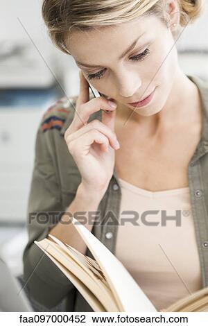 Stock Image - Woman using cell phone while reading book. Fotosearch