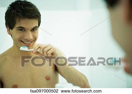 Stock Image - Young man looking in mirror, holding toothbrush, smiling at camera. Fotosearch