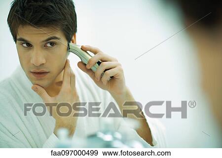 Stock Photo - Young man looking in mirror, shaving with electric razor. Fotosearch