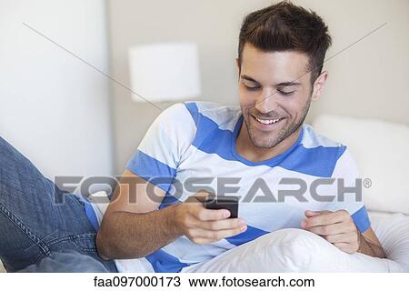 Young man reclining on bed looking at smartphone View Large Photo Image Stock Image - Young man reclining on bed looking at smartphone. Fotosearch