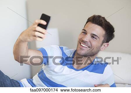 Young man reclining on bed taking selfie with smartphone View Large Photo Image Picture - Young man reclining on bed taking selfie with smartphone. Fotosearch