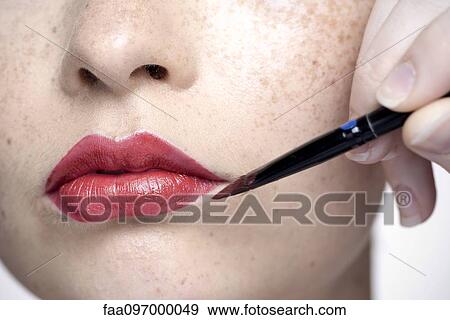 Stock Photo - Young woman applying lipstick with lip brush, close-up. Fotosearch