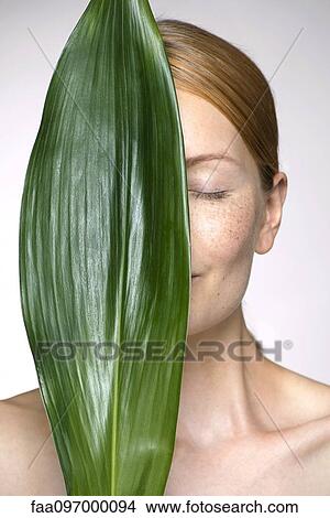 Picture - Young woman holding up leaf concealing part of face. Fotosearch