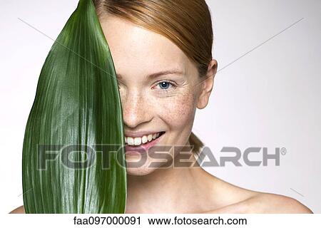 Stock Image - Young woman holding up leaf concealing part of face. Fotosearch