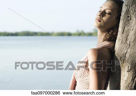 Young woman leaning against tree, eyes closed, lake in background View Large Photo Image Stock Photograph - Young woman leaning against tree, eyes closed, lake in background. Fotosearch