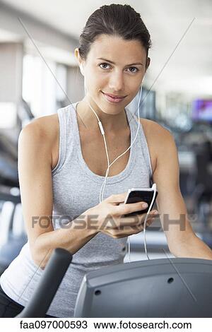 Stock Image - Young woman listening to music while using step climber at gym. Fotosearch