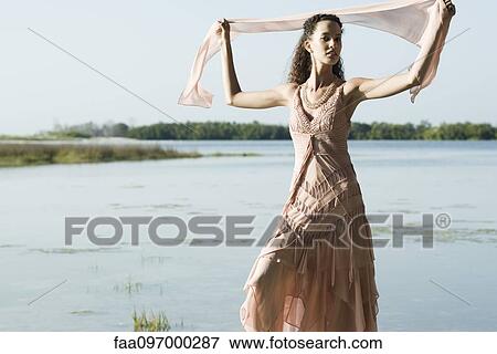 Stock Photo - Young woman wearing dress, holding up scarf, lake in background. Fotosearch