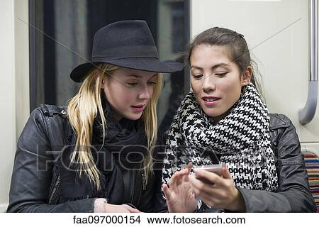 Young women sitting together on subway train, looking at smartphone View Large Photo Image Picture - Young women sitting together on subway train, looking at smartphone. Fotosearch