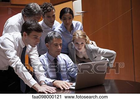 Stock Image - Business associates crowd around colleague looking over his shoulder at laptop computer. Fotosearch