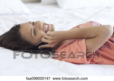 Stock Photo - Woman lying on bed chatting on cell phone. Fotosearch