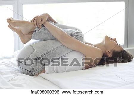 Stock Photograph - Woman lying on bed, hugging knees contentedly. Fotosearch