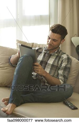 Stock Photo - Young man relaxing on couch at home using digital tablet. Fotosearch