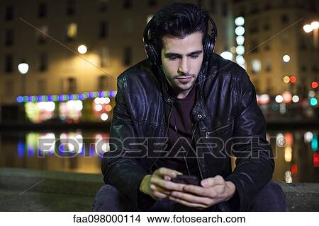Young man sitting alone outdoors at night listening to music with headphones View Large Photo Image Picture - Young man sitting alone outdoors at night listening to music with headphones. Fotosearch