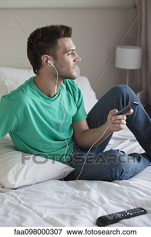 Young man sitting on bed listening to music with smartphone View Large Photo Image Stock Photo - Young man sitting on bed listening to music with smartphone. Fotosearch