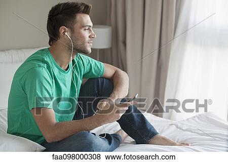 Stock Photo - Young man sitting on bed listening to music with smartphone. Fotosearch