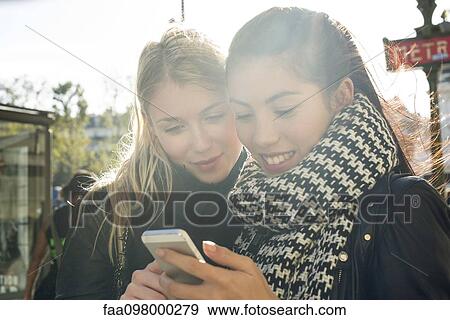 Stock Photo - Young women outdoors looking at cell phone together. Fotosearch