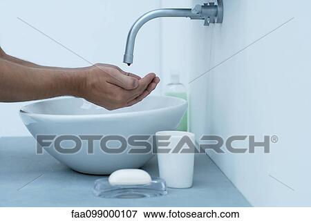 Stock Photo - Man holding hands under dripping faucet in bathroom sink, cropped. Fotosearch