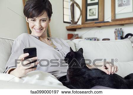 Stock Photo - Woman sitting on sofa with cat on her lap, using smartphone. Fotosearch