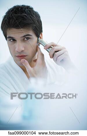 Picture - Young man looking in mirror, shaving with electric razor. Fotosearch