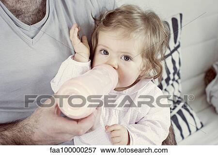 Stock Photo - Baby girl sitting on father's lap, drinking from bottle, cropped. Fotosearch
