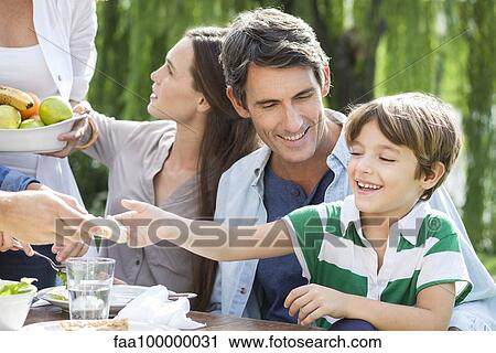 Stock Image - Family eating healthy meal together outdoors, father holding son on lap. Fotosearch