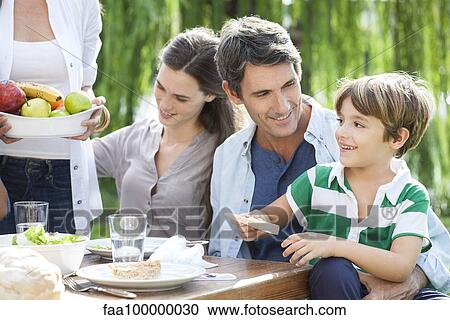 Stock Image - Family eating together outdoors, father holding son on lap. Fotosearch