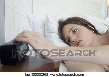 Stock Photo - Woman in bed pressing alarm clock snooze button. Fotosearch